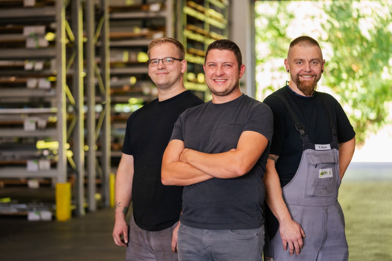 Drei Männer in Arbeitskleidung stehen lächelnd vor Lagerregalen mit Metallprofilen.