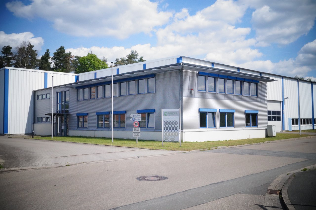 Modernes Bürogebäude mit blauen Fensterrahmen und angrenzender Lagerhalle unter bewölktem Himmel.