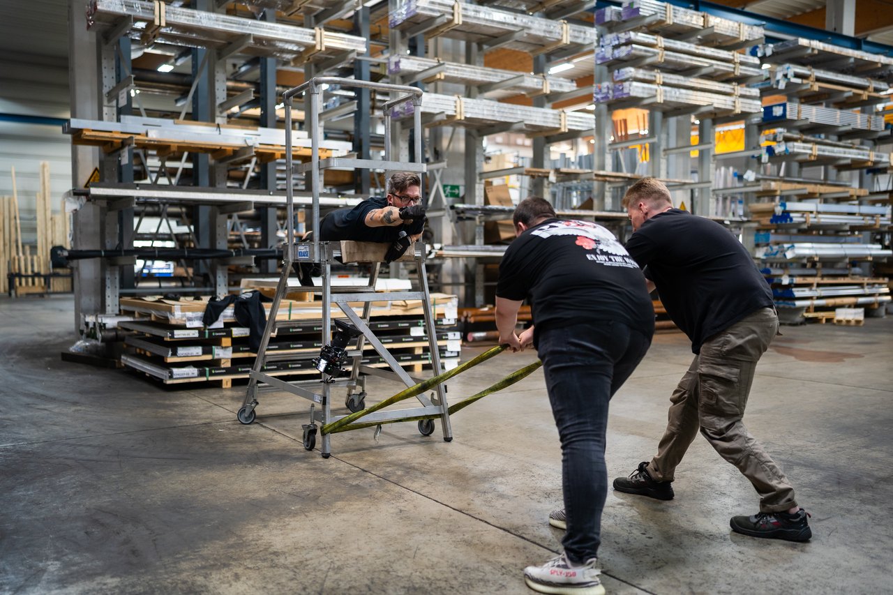 Zwei Männer ziehen einen dritten Mann, der auf einer Leiter liegt, durch eine Lagerhalle mit Metallregalen.